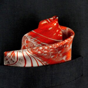 May include: A red and white patterned pocket square with a paisley design. The pocket square is folded and placed in a black pocket.