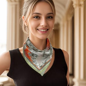 May include: A square silk scarf with a detailed floral and foliage pattern in shades of gray, orange, and green. The scarf is worn around the neck and has a satin finish. The background is blurred, with a woman wearing a black top.