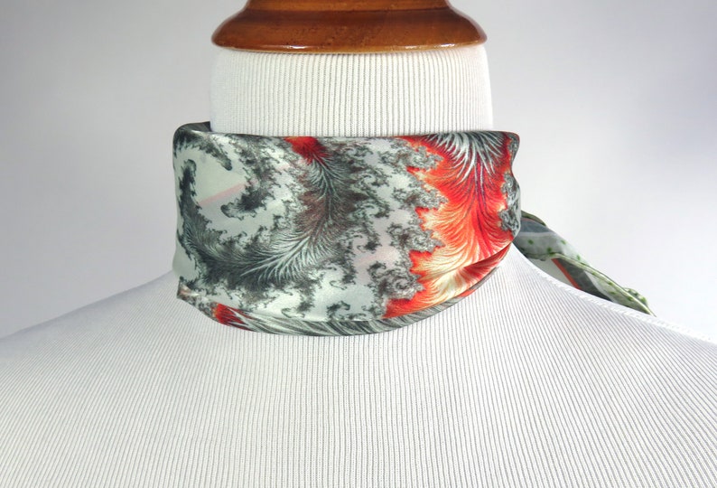May include: A silk scarf with a unique pattern of gray, red, and white feather-like designs. The scarf is wrapped around a mannequin's neck, showcasing its vibrant colors and intricate details. The scarf's design is eye-catching and adds a touch of elegance.