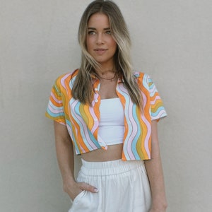 May include: A woman wearing a cropped, short-sleeved shirt with a colorful wavy pattern. The shirt has a button-up front and is paired with white high-waisted pants.