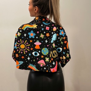 May include: Black cropped jacket with a colorful, whimsical print featuring rainbows, planets, mushrooms, and stars. The jacket has a collared neckline and short sleeves. The design is set against a black background.