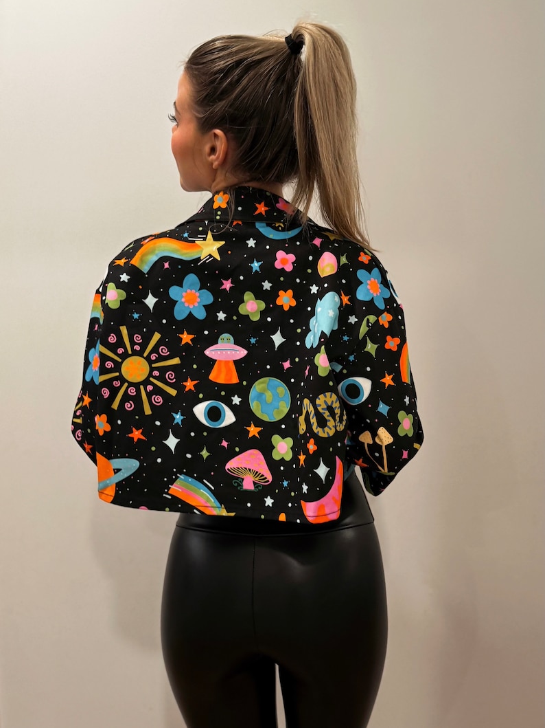 May include: Black cropped jacket with a colorful, whimsical print featuring rainbows, planets, mushrooms, and stars. The jacket has a collared neckline and short sleeves. The design is set against a black background.