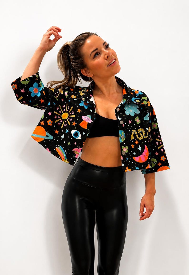 May include: Black button-up shirt with a colorful space-themed pattern featuring planets, stars, and UFOs. The shirt has a white collar and cuffs. The model is wearing black leggings and a black sports bra.