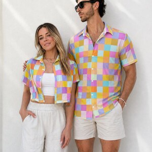 May include: Two short-sleeved button-down shirts featuring a vibrant, multi-colored checkered pattern in shades of pink, yellow, light blue, and mustard. The shirts have a classic collar and a front button closure, showcasing a repeating square grid design.