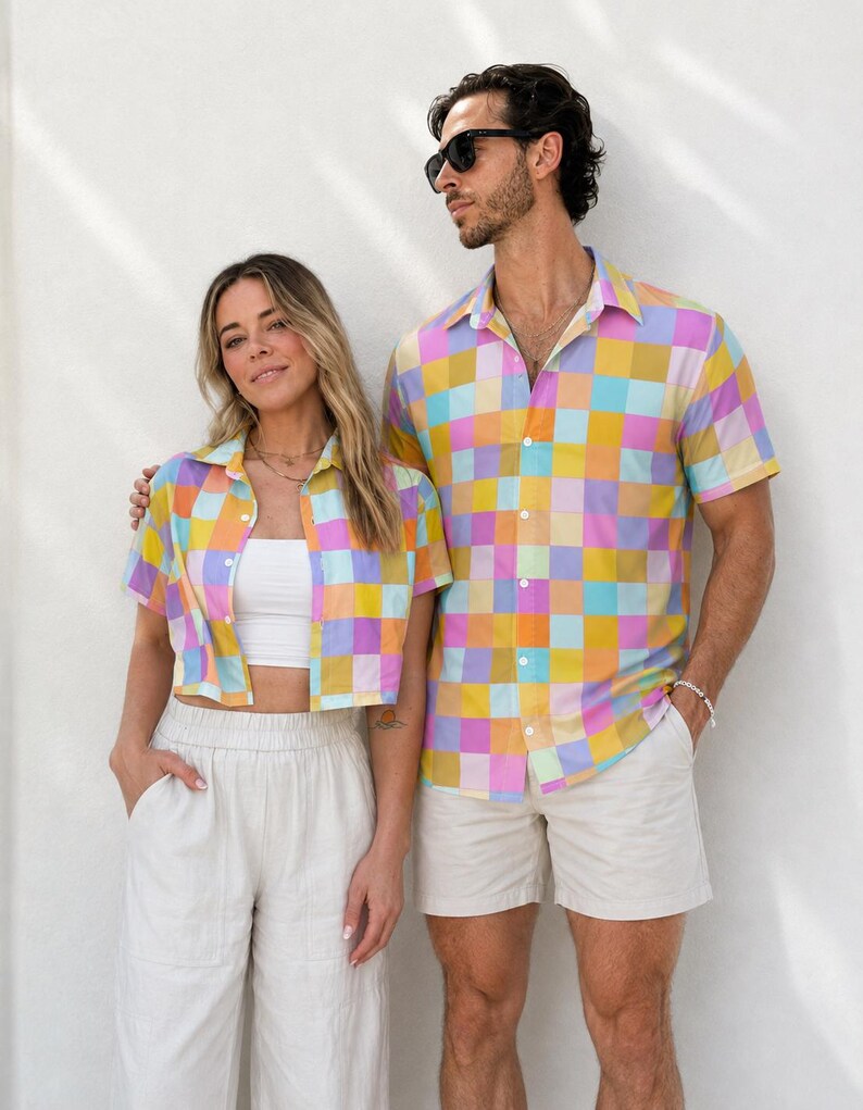 May include: Two short-sleeved button-down shirts featuring a vibrant, multi-colored checkered pattern in shades of pink, yellow, light blue, and mustard. The shirts have a classic collar and a front button closure, showcasing a repeating square grid design.