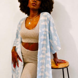 May include: A light blue open-front robe featuring a repeating white cloud pattern, worn over a beige fuzzy cropped tank top and matching high-waisted leggings. The robe has wide sleeves and falls to a mid-calf length, creating a layered ensemble.