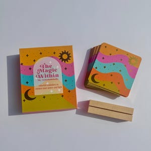 May include: A deck of "The Magic Within" cards with a matching box and wooden stand. The box and card backs feature a colorful design with a sun, moon, stars, and wavy lines. The box reads "colorful reminders to awaken your power and light."