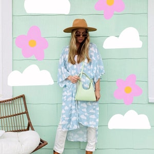 Puede incluir: Una mujer lleva un kimono azul claro con estampado de nubes blancas, pantalones blancos, botas marrones y un bolso de mano verde claro. Está de pie frente a una pared verde claro con flores rosas y nubes blancas.