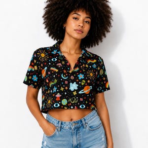 May include: A black short-sleeved button-down crop top featuring a colorful all-over print of planets, stars, rainbows, flowers, mushrooms, and alien faces, paired with light blue denim shorts that have a distressed, frayed hemline.