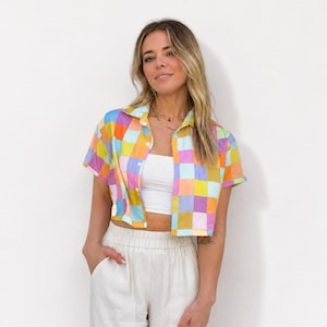 May include: A short-sleeved button-down shirt featuring a multicolored checkered pattern in shades of yellow, pink, blue, and orange. The shirt is worn open over a plain white tube top, paired with light beige high-waisted trousers.