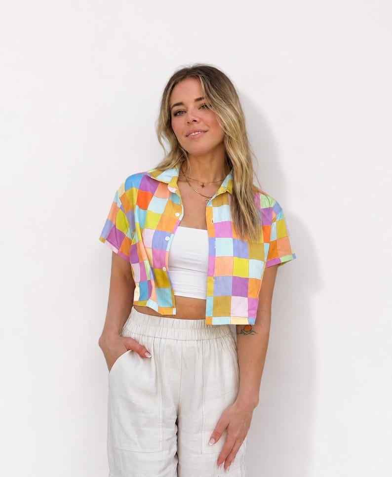 May include: A short-sleeved button-down shirt featuring a multicolored checkered pattern in shades of yellow, pink, blue, and orange. The shirt is worn open over a plain white tube top, paired with light beige high-waisted trousers.