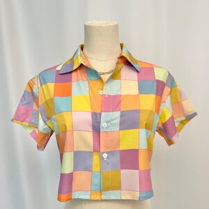 May include: A short-sleeved button-down shirt featuring a multi-colored checkered pattern in shades of yellow, orange, pink, light blue, and lavender. The garment has a classic collar, a straight hemline, and three white buttons down the front.