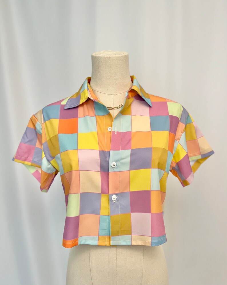 May include: A short-sleeved button-down shirt featuring a multi-colored checkered pattern in shades of yellow, orange, pink, light blue, and lavender. The garment has a classic collar, a straight hemline, and three white buttons down the front.