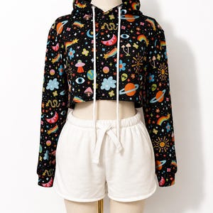 May include: A black cropped hoodie featuring a colorful space-themed print with planets, stars, rainbows, mushrooms, and alien motifs, paired with plain white drawstring shorts. The hoodie includes a hood with long white drawstrings and ribbed cuffs.