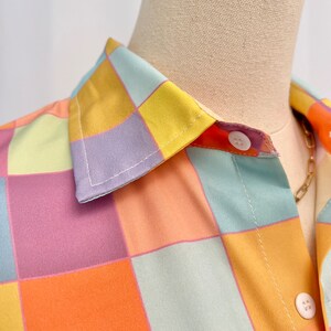 May include: A button-down shirt featuring a repeating checkered pattern in pastel shades of yellow, orange, light blue, and lavender. The garment has a classic pointed collar and white circular buttons along the front placket.