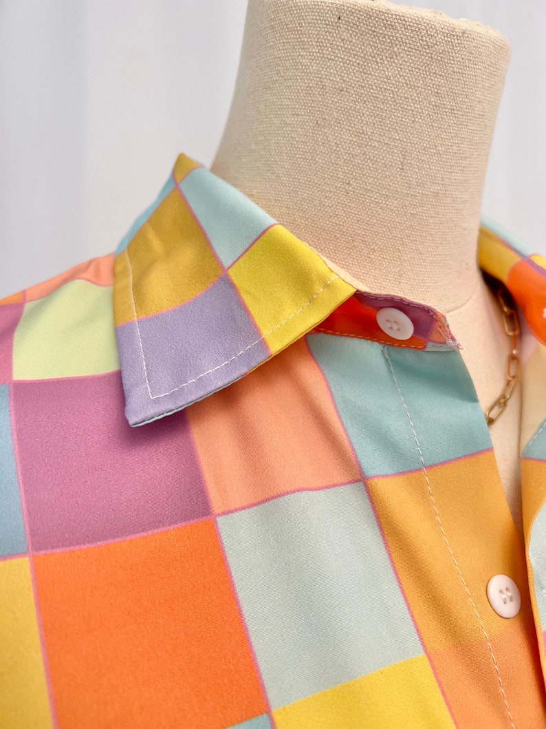 May include: A button-down shirt featuring a repeating checkered pattern in pastel shades of yellow, orange, light blue, and lavender. The garment has a classic pointed collar and white circular buttons along the front placket.