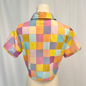 May include: A short-sleeved button-down shirt featuring a repeating checkered pattern in pastel shades of yellow, orange, light blue, and lavender. The garment has a classic collar and a straight hemline, displayed on a mannequin torso.