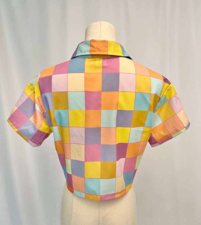 May include: A short-sleeved button-down shirt featuring a repeating checkered pattern in pastel shades of yellow, orange, light blue, and lavender. The garment has a classic collar and a straight hemline, displayed on a mannequin torso.