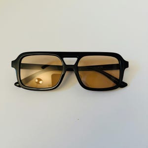 May include: Black square-framed sunglasses with brown tinted lenses. The sunglasses have a double bridge design and a thick frame. The lenses are a warm brown color, and the frame is a glossy black.
