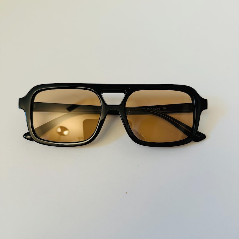 May include: Black square-framed sunglasses with brown tinted lenses. The sunglasses have a double bridge design and a thick frame. The lenses are a warm brown color, and the frame is a glossy black.