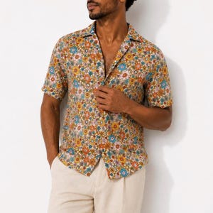 May include: A short-sleeved button-down shirt featuring a dense, colorful floral pattern with orange, blue, pink, and yellow flowers on a tan background. The shirt has a classic collar, a front button placket, and is paired with light beige trousers.