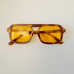 May include: Brown square-framed sunglasses with yellow lenses. The sunglasses have a classic, retro design with a keyhole bridge and thick temples. The frames have a tortoiseshell pattern.