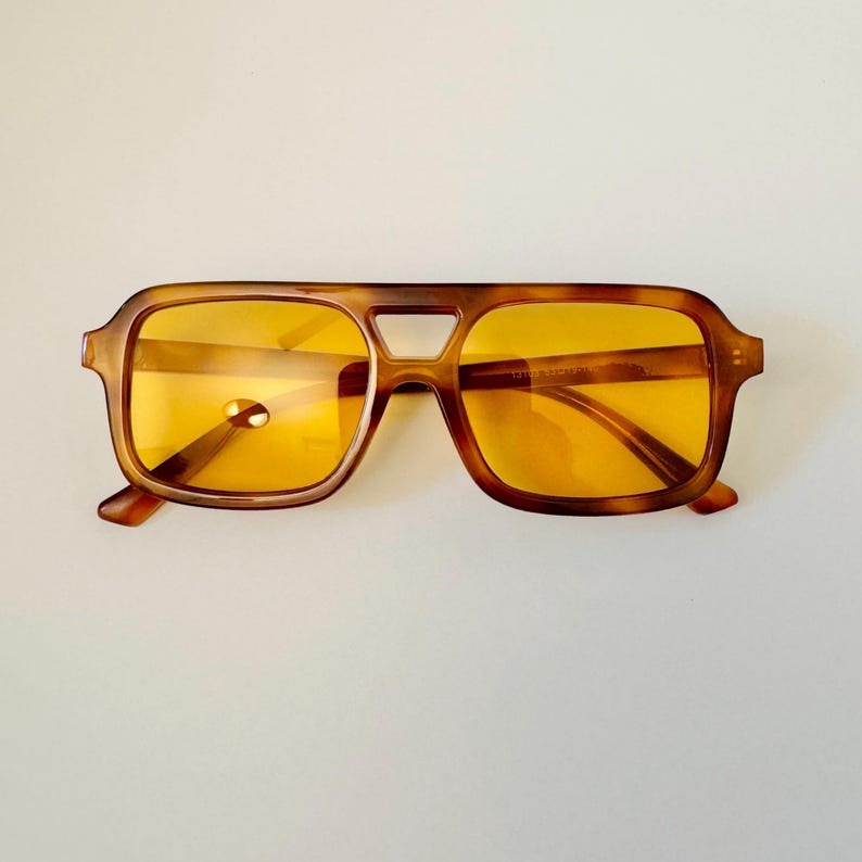 May include: Brown square-framed sunglasses with yellow lenses. The sunglasses have a classic, retro design with a keyhole bridge and thick temples. The frames have a tortoiseshell pattern.