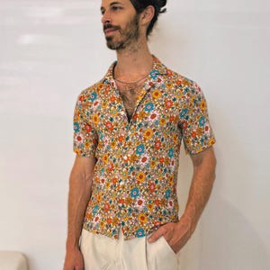 May include: Short-sleeved button-down shirt with a vibrant floral pattern in orange, yellow, blue, and pink. The shirt is paired with light beige pants. The shirt's collar is open, and the overall style is casual.