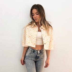 May include: A cream-colored, short-sleeved jacket with a celestial pattern of moons and stars. The jacket has a button-up front and is paired with light-wash denim jeans. The outfit is casual and stylish.