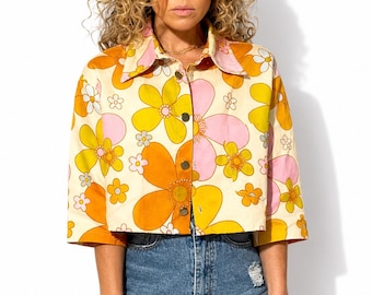 Puede incluir: Una camisa corta abotonada con fondo crema y un estampado floral a gran escala en tonos naranja, amarillo y rosa. La camisa tiene cuello en punta, mangas tres cuartos y un dobladillo recto, combinada con unos pantalones cortos de mezclilla azul.