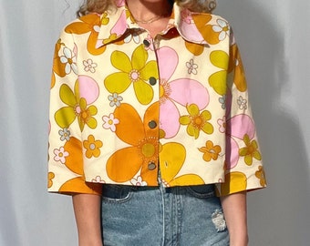 May include: A cropped, cream-colored shirt with a retro floral print in shades of pink, orange, and green. The shirt has a button-up front and short sleeves.