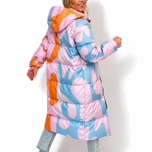 May include: A long, quilted puffer coat featuring a hooded design and a vibrant, abstract pattern in shades of light pink, bright orange, and sky blue. The coat has a full-length silhouette with horizontal stitching and long sleeves with cuffs.