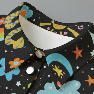 May include: Close-up of a black shirt collar with a whimsical pattern. The design features a yellow snake, rainbows, flowers, stars, and clouds in various colors. The shirt has two silver buttons.