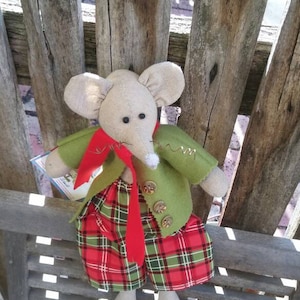 May include: A plush mouse toy wearing a green jacket, red scarf, and plaid skirt. The mouse is sitting on a wooden bench with a slatted back.