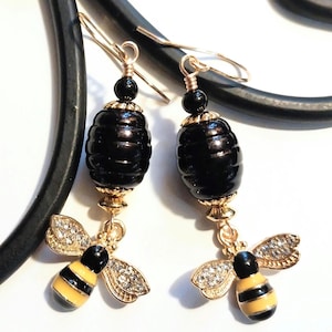 May include: A pair of gold-toned earrings featuring black, barrel-shaped beads and bee charms. The bee charms have yellow and black striped bodies, with rhinestone-studded wings. The earrings hang from gold-colored ear wires.