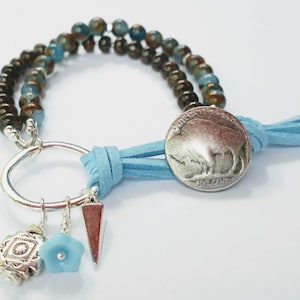 May include: A blue and brown beaded bracelet with a silver charm featuring a buffalo nickel, a silver flower charm, and a silver triangle charm. The bracelet is tied with a blue leather cord.