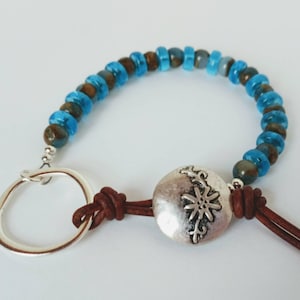 May include: A handmade bracelet featuring a mix of blue and brown beads, a silver-tone metal ring, and a decorative silver-tone metal disc with a floral design. The bracelet is connected with brown leather cords.