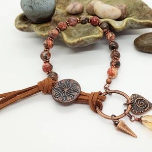 May include: A handcrafted bracelet featuring speckled red and brown beads, copper accents, and a suede leather tassel. The bracelet includes a decorative button and charms, including a heart and a yellow teardrop-shaped bead.
