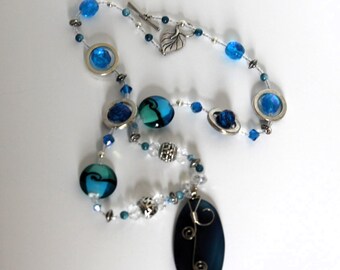 Può includere: Una collana con un grande ciondolo ovale blu scuro con spirali in filo d'argento, impreziosita da perle di vetro blu e turchese, anelli in metallo argentato, cristalli sfaccettati trasparenti e un ciondolo a forma di foglia d'argento.
