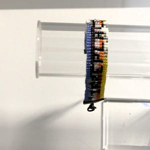 Pu&ograve; includere: Un braccialetto di perline multicolori con una chiusura argentata. Il braccialetto presenta un motivo di perline blu, bianche, arancioni e gialle.