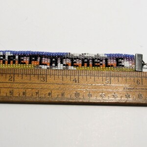 Pu&ograve; includere: Un braccialetto di perline con un design colorato e la parola "IRREPLACEABLE" in lettere nere. Il braccialetto viene misurato con un righello di legno, mostrando che &egrave; lungo circa 15 cm.