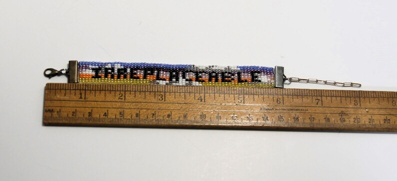 Pu&ograve; includere: Un braccialetto di perline con un design colorato e la parola "IRREPLACEABLE" in lettere nere. Il braccialetto viene misurato con un righello di legno, mostrando che &egrave; lungo circa 15 cm.