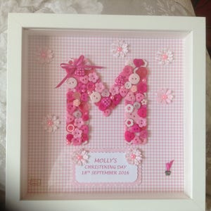 Puede incluir: Un marco blanco con un fondo de cuadros vichy rosa y blanco. La letra M está hecha de botones rosas y un lazo rosa. El texto "Molly's Christening Day 18th September 2016" está en una tarjeta rosa.