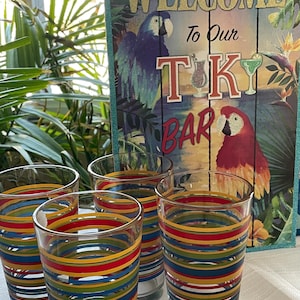 Può includere: Quattro bicchieri trasparenti con strisce orizzontali di vari colori, tra cui blu, rosso, giallo e verde. Un cartello di legno con la scritta "WELCOME To Our TIKI BAR" è sullo sfondo.