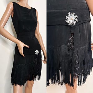 May include: Black sleeveless dress with a fitted bodice and a fringed skirt. The dress features a decorative brooch at the waist and a lace panel. The dress is a vintage style.