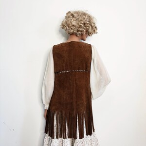 May include: A brown suede vest with fringe detail, worn over a white blouse with long sleeves and a floral print dress. The vest has a V-neck and a decorative stitched design.