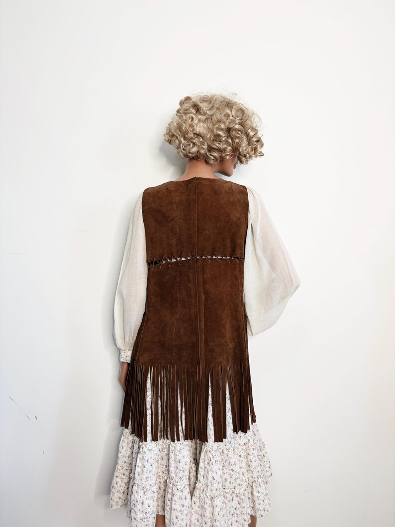 May include: A brown suede vest with fringe detail, worn over a white blouse with long sleeves and a floral print dress. The vest has a V-neck and a decorative stitched design.