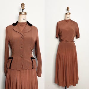 Puede incluir: Un traje vintage de dos piezas y un vestido en un tono marrón rojizo apagado. El traje presenta una chaqueta con cuello con ribete negro y botones, combinada con una falda plisada. El vestido tiene cuello alto, mangas cortas y cinturón a juego.