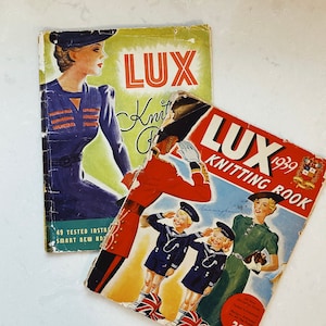 Peut inclure: Deux magazines vintage "LUX Knitting Book". L'un présente une femme en robe bleue, l'autre un soldat et des enfants en marins. Les deux magazines sont usés, avec des couleurs fanées et des signes de vieillissement.