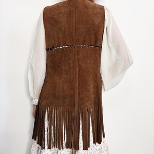 May include: A brown suede vest with long fringe at the hem. The vest has a stitched design on the back and is worn over a white blouse with billowing sleeves and a floral print dress.